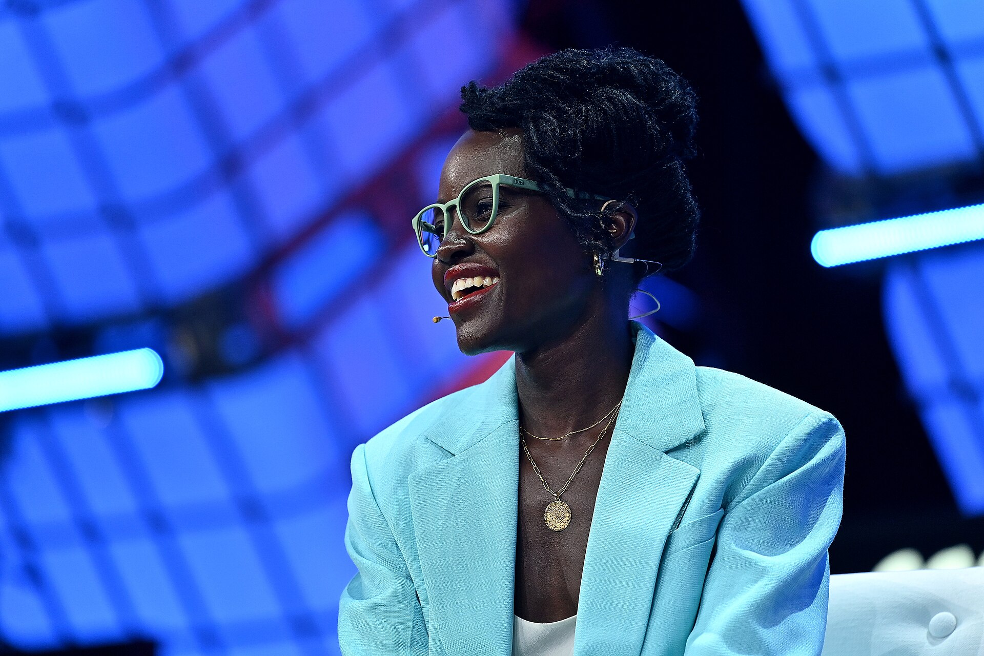 Lupita Nyong'o speaks on a conference stage in a light blue blazer and green glasses, smiling, wearing a headset microphone.